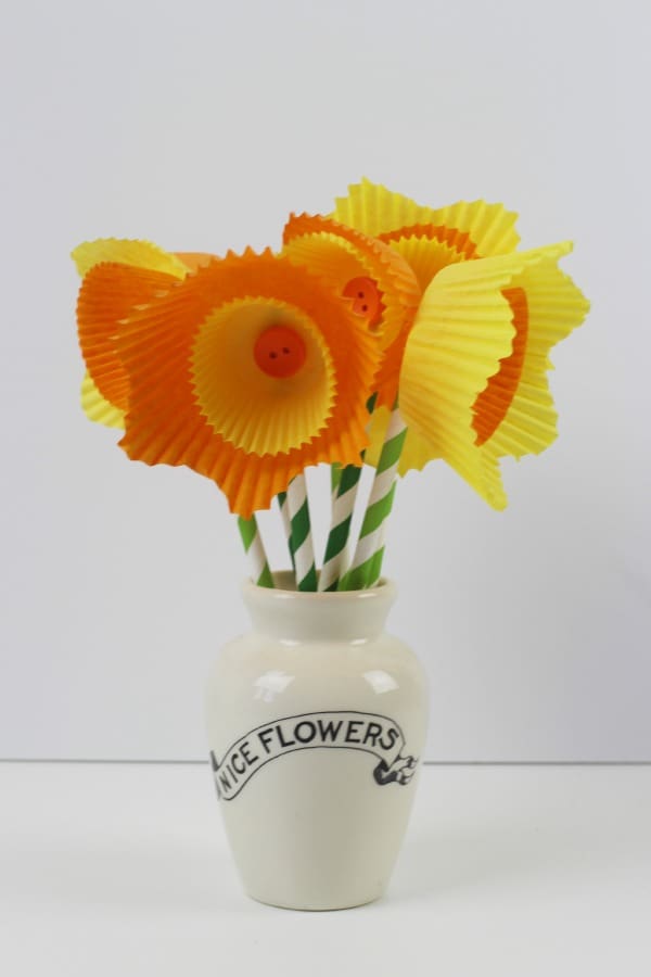 A number of yellow and orange cake cases layered to look like daffodil flowers with buttons in the center and paper straws as stems