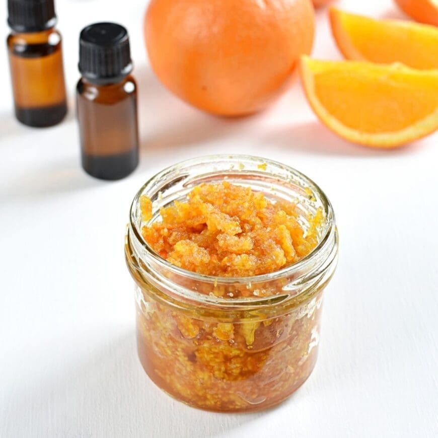 A jar of homemade salt scrub with two bottles of essential oil in the background and orange segments