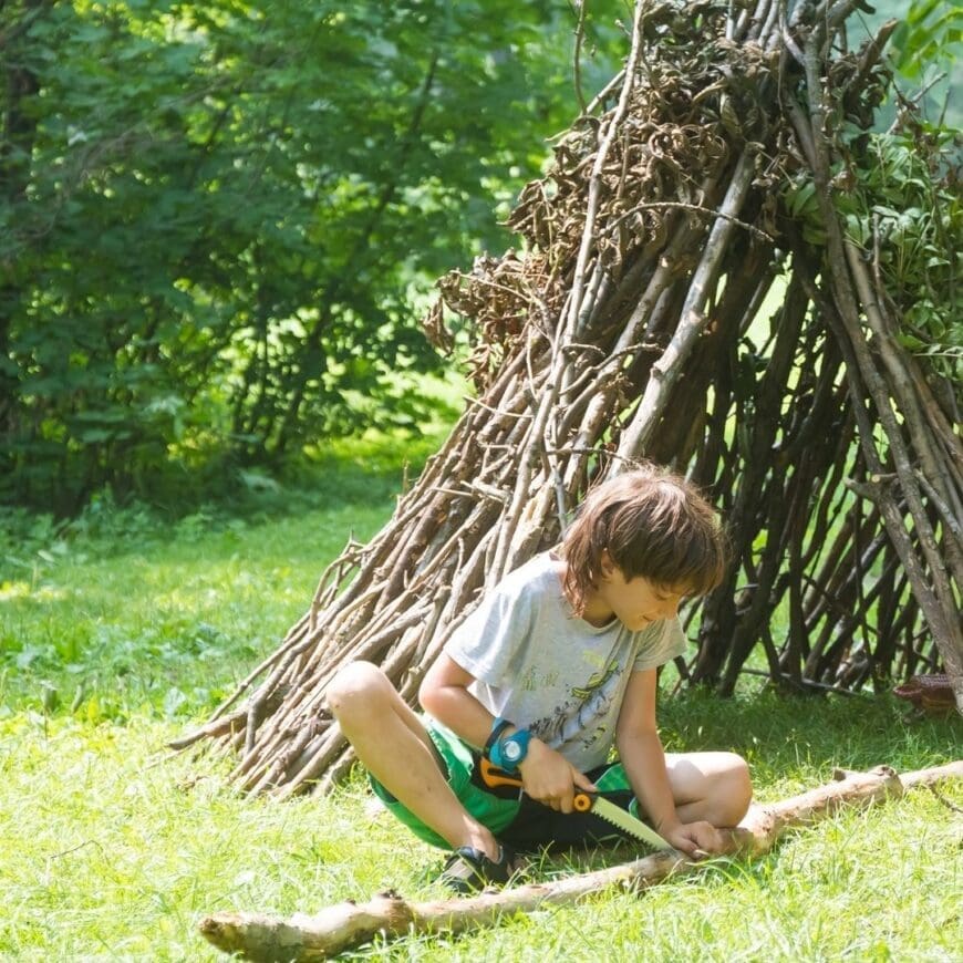 Young boy cutting branches with a hand saw in front of a tent structure made from twigs, branches and leaves