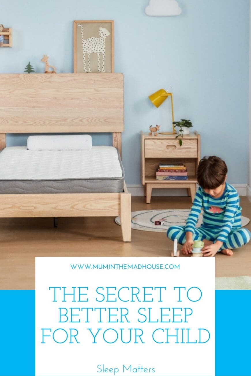 A young child in blue-striped pajamas sleeping soundly on a white mattress in a cozy bedroom with a wooden bed frame. The room has soft blue walls, wooden toys, and a framed artwork above the bed, creating a calming sleep environmen