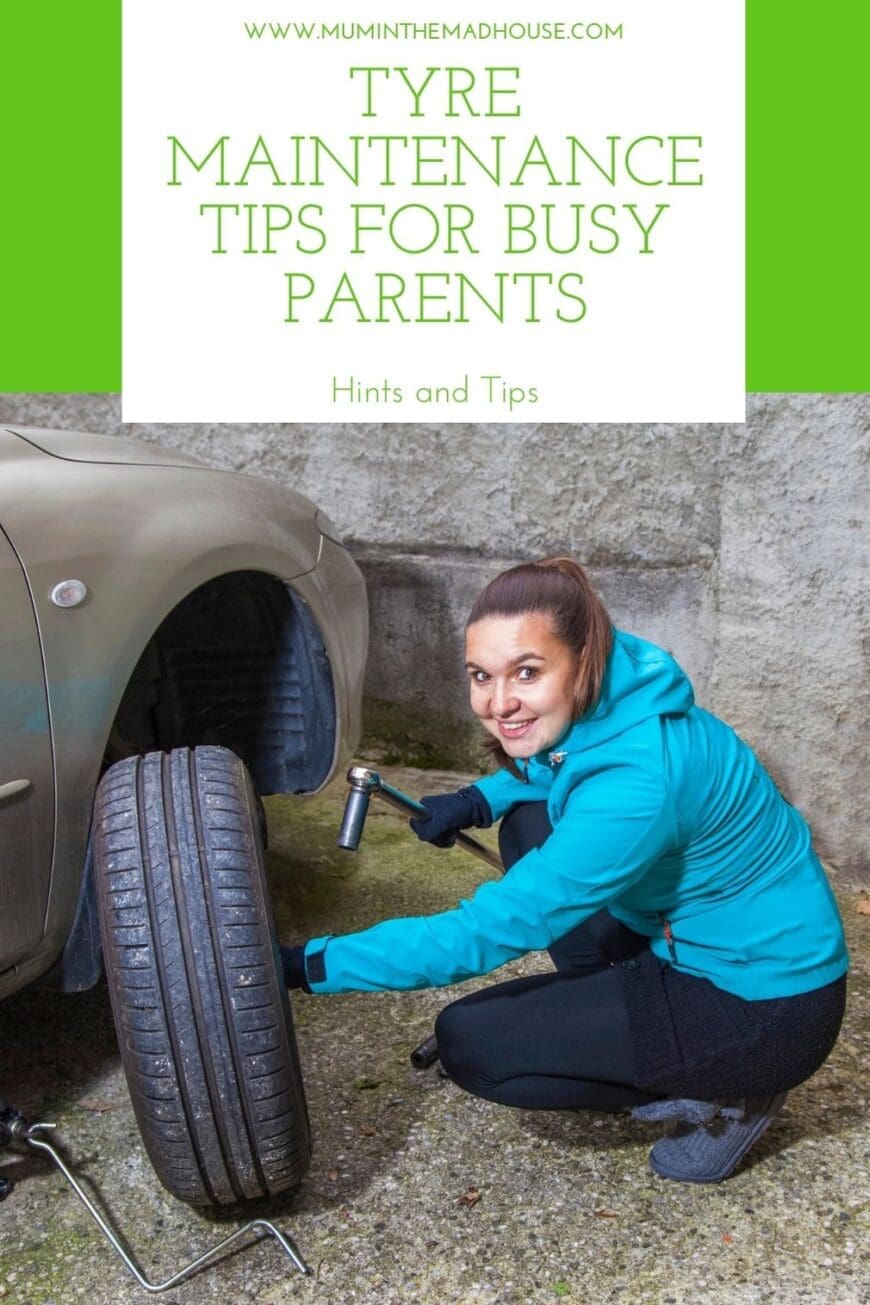 A smiling woman in a blue jacket kneels beside a car while changing a tyre. She holds a wrench and looks confident as she works on the vehicle, demonstrating essential tyre maintenance skills for busy parents