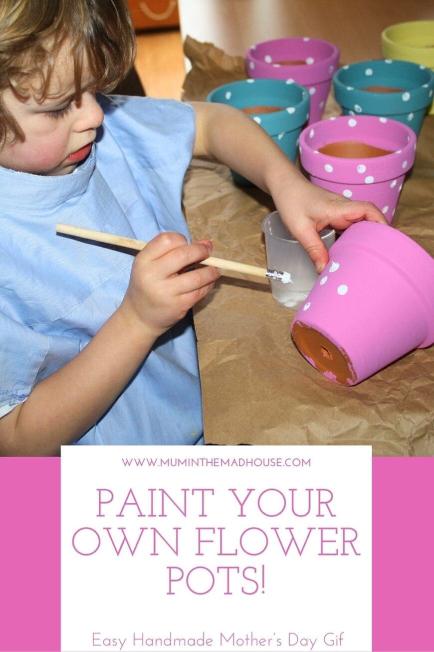 A young child in a blue apron is painting a pink terracotta flower pot with white polka dots, using a pencil eraser as a stamping tool. Several painted pots in pink, blue, and yellow are drying in the background. The text overlay reads “Paint Your Own Flower Pots! – Easy Handmade Mother’s Day Gift” from Mum in the Mad House.